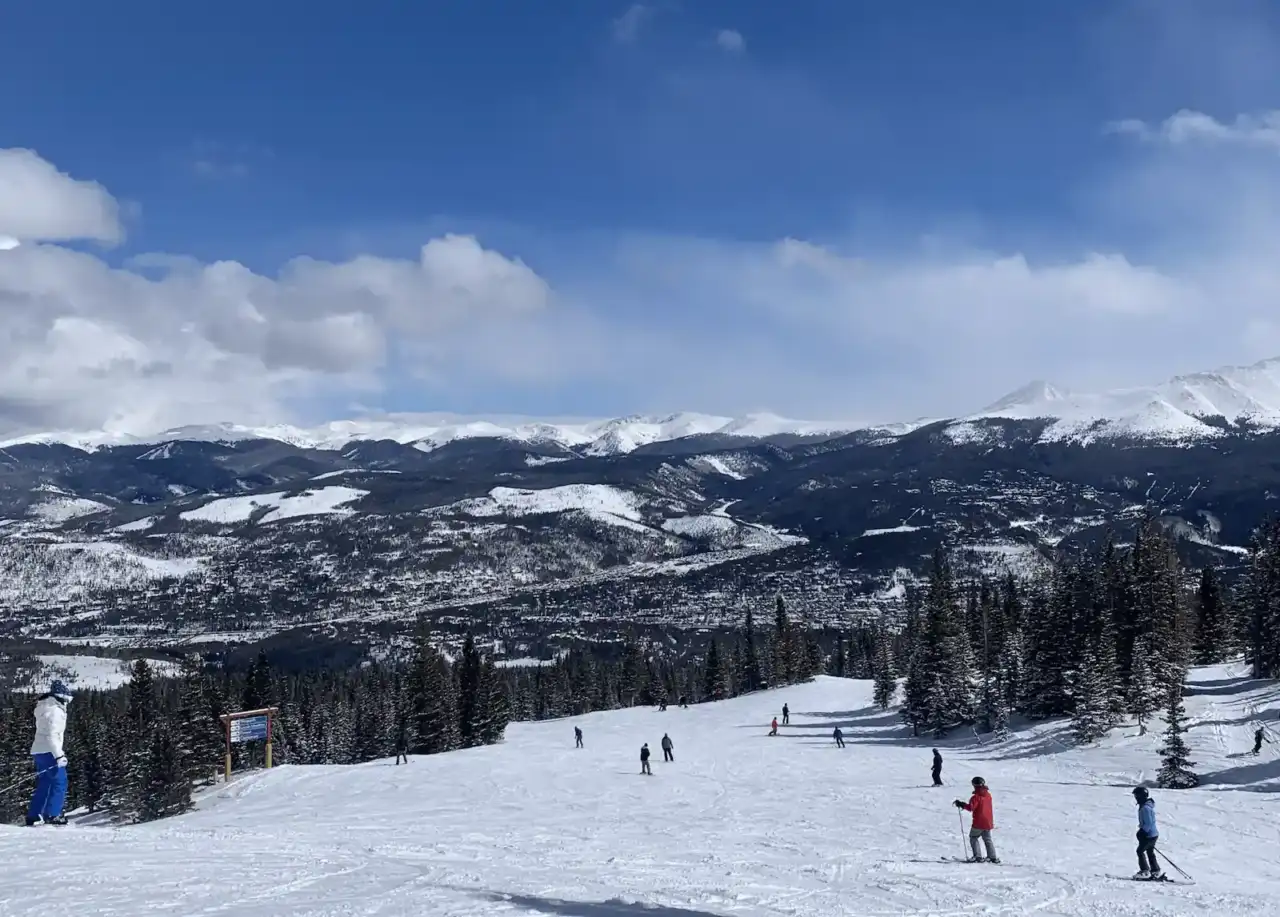 Scenic winter photograph of a ski resort with skiers on snowy slopes, snow-capped Rocky Mountains in the background under blue sky with white clouds. Breckenridge Ski Resort