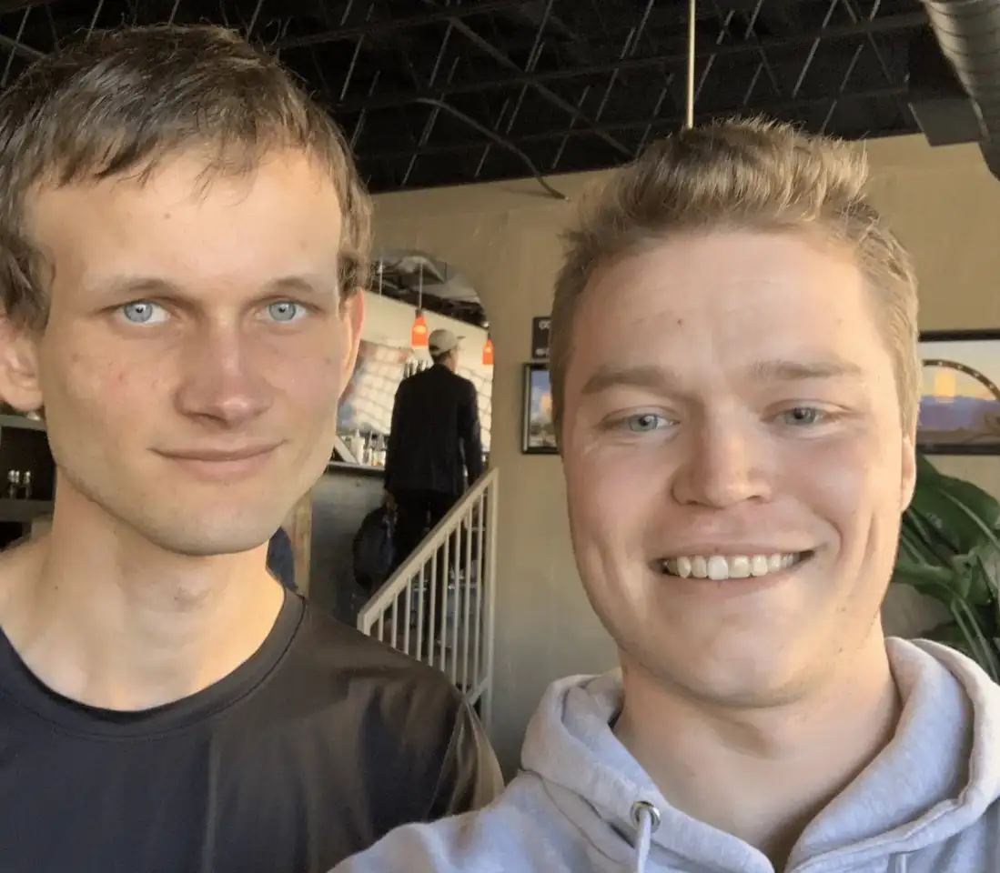 Selfie photo of Ethereum co-founder Vitalik Buterin with another man in a casual indoor setting, Buterin wearing a dark t-shirt and the other man wearing a gray hoodie.