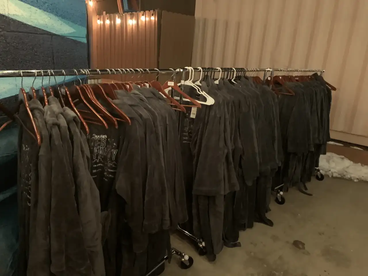 Clothing racks with dark gray hoodies and t-shirts on wooden hangers at a Web3 event venue, with string lights visible in the background.