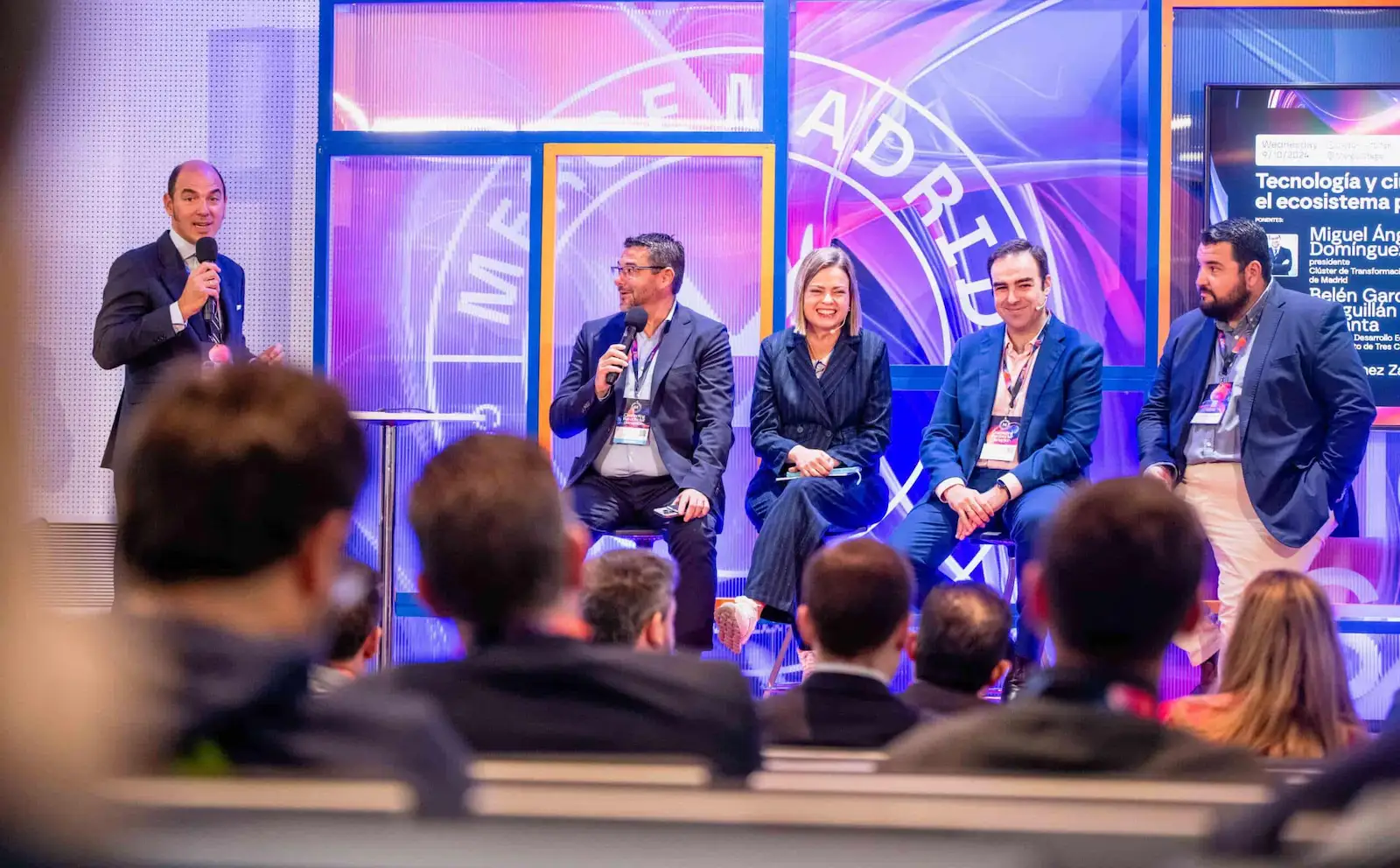 Technology conference panel discussion in Madrid with five speakers on stage, colorful backdrop with "MADRID" text, discussing technology and ecosystem topics in Spanish.