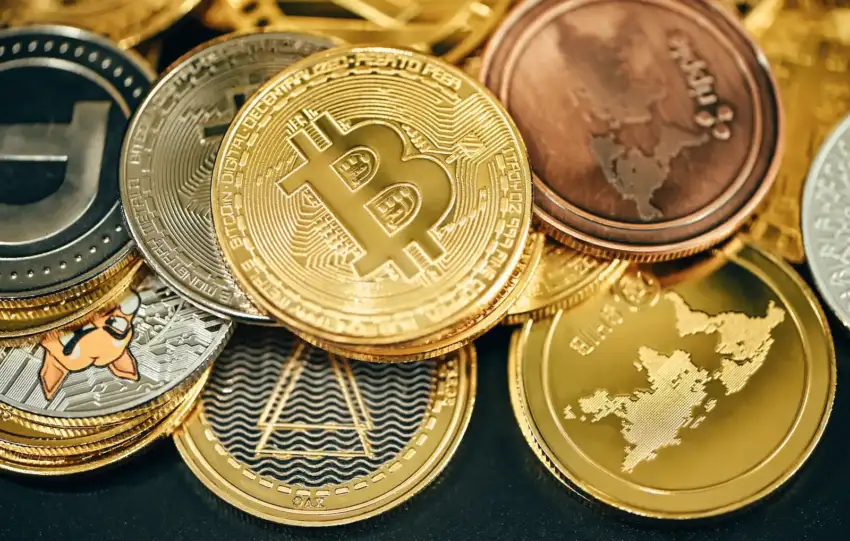 Close-up photograph of various physical cryptocurrency coins piled together, featuring a prominent gold Bitcoin in the center, surrounded by Dogecoin, Litecoin, Ripple, and other altcoin tokens on a dark surface.