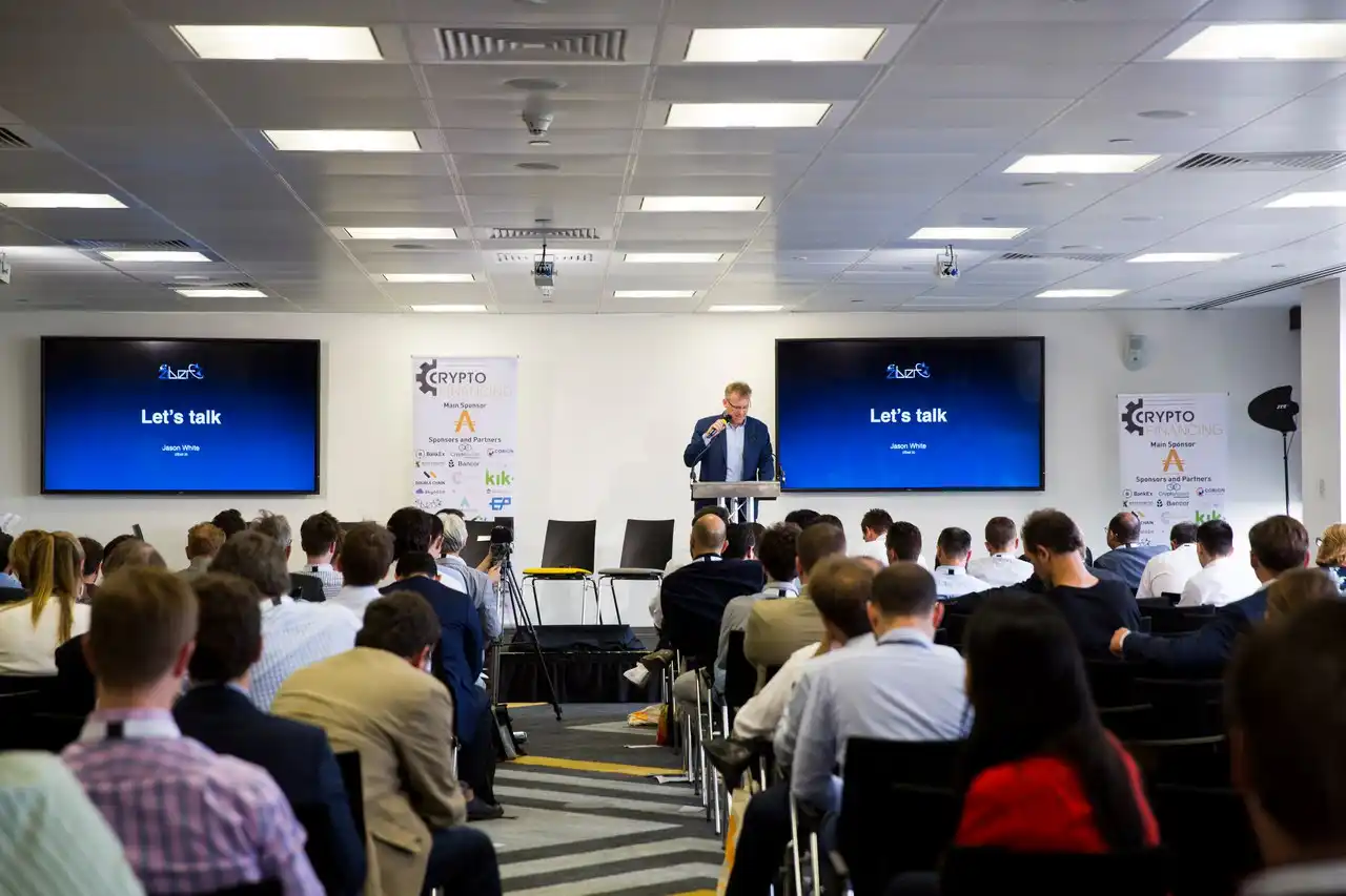 Jason White at podium addressing audience at CryptoFinancing 2017 conference in London, with dual screens showing Ziber logo and "Let's talk - Jason White" presentation slides, sponsor banners visible