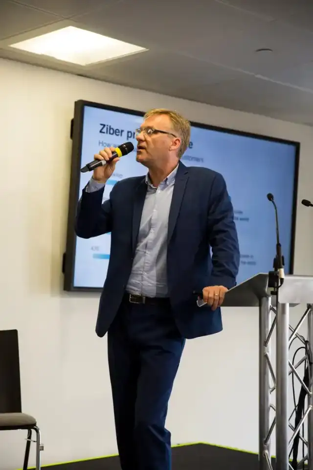 Ziber Operations lead Jason White speaking into microphone during presentation with slide showing Ziber platform details and iOS reference