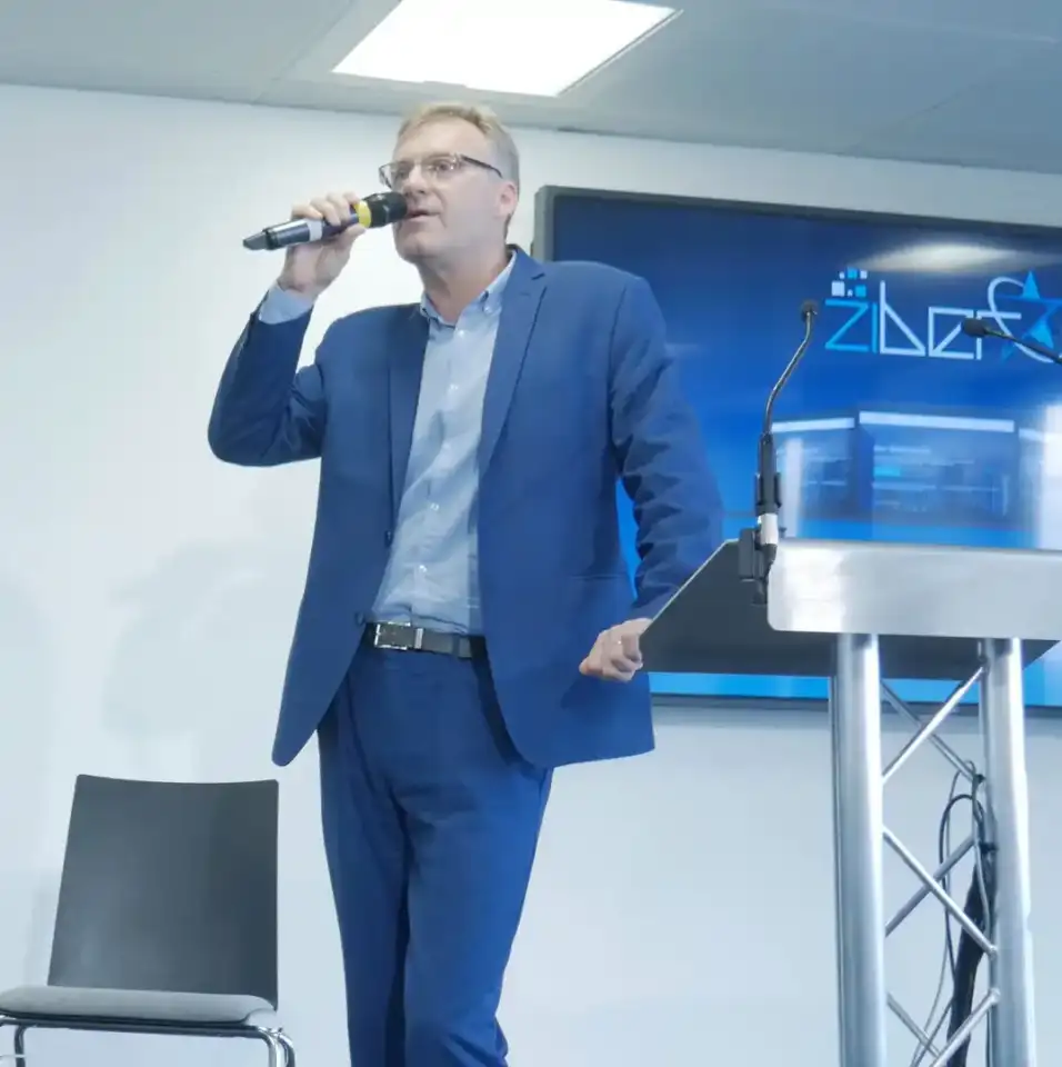 Ziber Operations lead Jason White speaking into microphone at podium during 2017 ICO launch event, wearing blue suit, with Ziber logo displayed on screen behind him