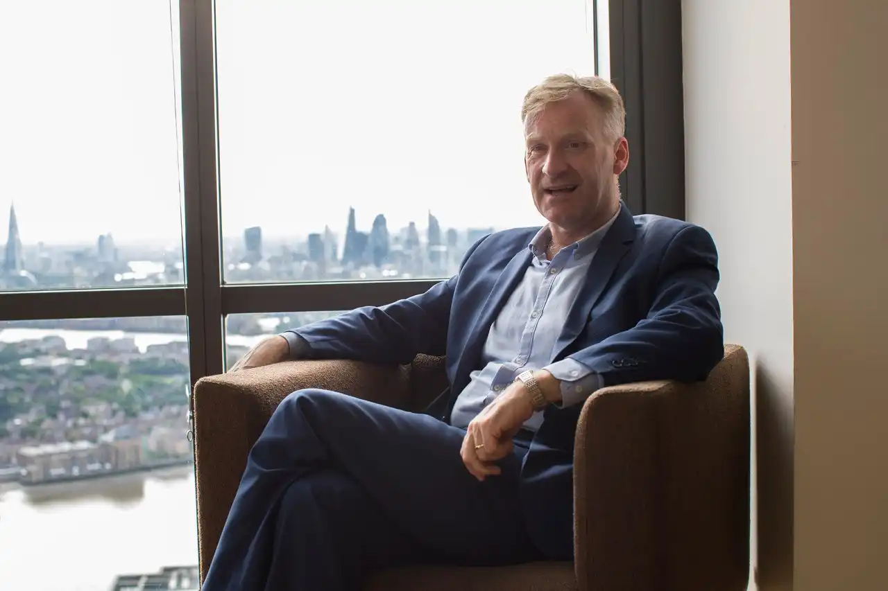 Ziber Operations lead Jason White seated in brown armchair by window overlooking London skyline including The Shard and Thames River, wearing blue suit
