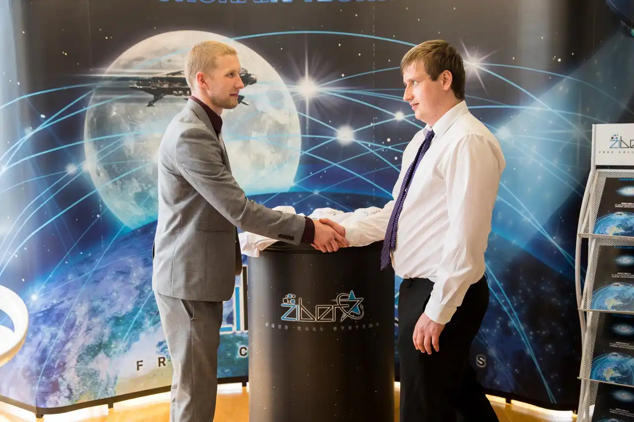 Ziber CEO Mark Braun in white shirt and purple tie shaking hands with associate in gray suit at Free Call Systems promotional booth with cosmic-themed backdrop featuring moon and Earth